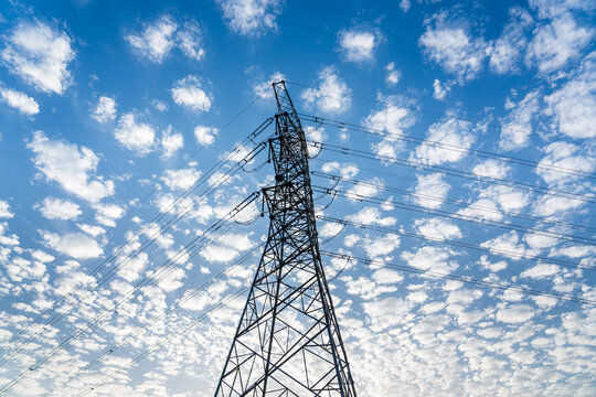 A low-angle photograph captures a steel lattice transmission tower against a scattered cloud filled bright blue sky, with overhead power lines stretching across the frame.