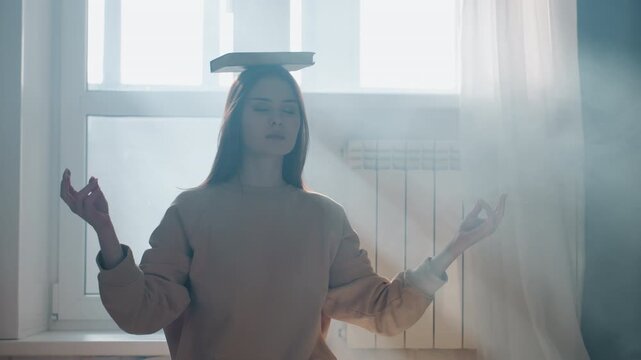 Young woman balancing book on head near window light practicing breathing technique as grammar coach exercise, palms raised, eyes open, soft curtain, misty rays, quiet study space, intentional