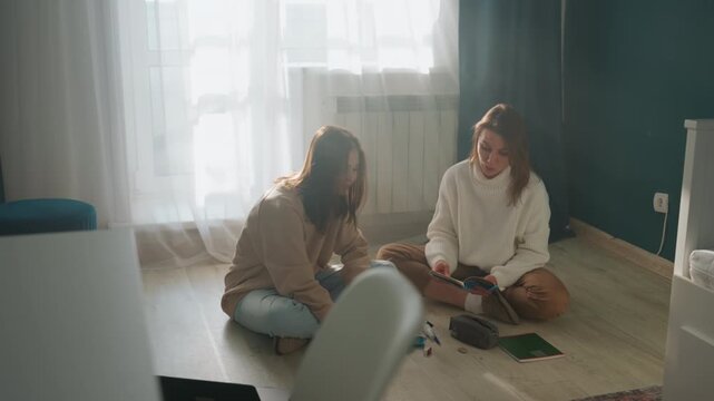 Two women studying on floor in sunlit living space with laptop, notebooks, pens and grammar workbook gentle tutor offering feedback, casual sweaters, warm natural light, calm supportive atmosphere