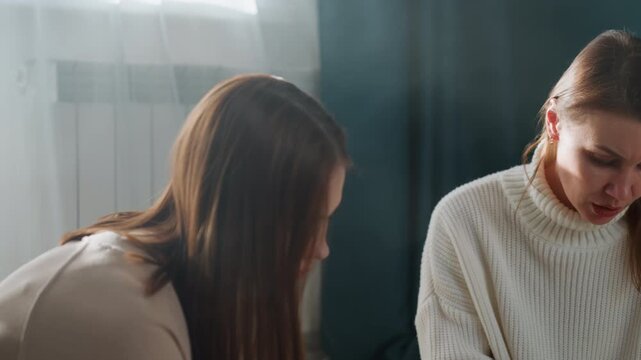 Two women proofreading grammar at home, tutor guiding student over handwritten notes at small table, knit sweaters with teal wall background, concentrated expressions and gentle dialogue, soft