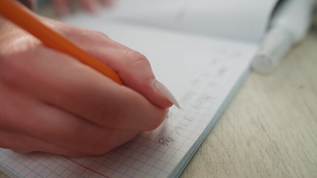 Closeup hand writing grammar exercise with orange pencil on grid paper, neat manicure visible, correction fluid near edge, focused concentration and steady strokes, solitary practice in home study