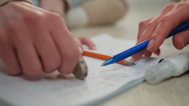 Closeup hands erasing pencil marks on workbook page careful correction during home study student practicing math exercises orange pencil visible soft daylight focused fingertips on grid sheet casual