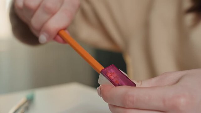 Caucasian hands sharpening pencil at home desk, tutor preparing grammar practice materials, close detail of orange pencil and purple sharpener, neat shavings falling onto white table, warm natural