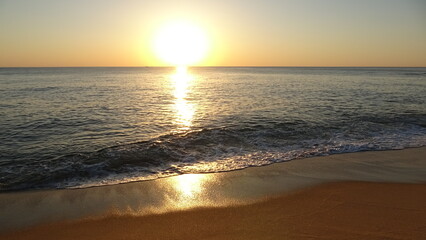Obraz premium Majestic Sunrise over East Sea Horizon. The bright sun is rising over the ocean horizon, casting a strong golden reflection. Gentle waves with foam wash onto the sandy foreground beach.