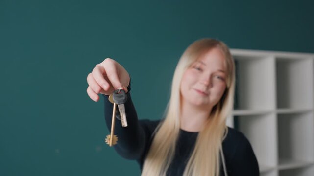 Caucasian woman holding new keys in empty apartment with teal wall and shelving, blonde hair in black sweater, smiling while offering keyring toward camera, celebratory homeowner marking movein
