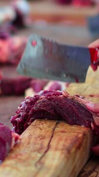 Hands cut raw meat on a wooden block. Fresh meat pieces lie around workspace.