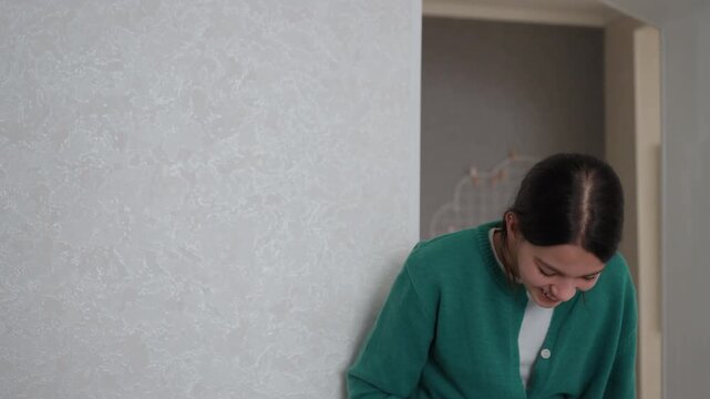 Young woman apartment smiling while holding potted plant, shy laughter and downward glance evolve into bright grin, green cardigan against textured wall, natural morning light, minimal interior, calm