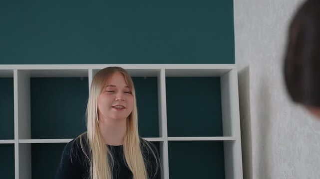 Laughing white woman with friend moving, teal shelving backdrop, brunette helper blurred foreground, boxes and packing, handshake and highfive, teamwork energy, cozy sweater candid conversation movein