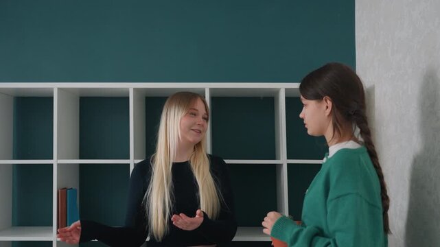 Two caucasian women empty apartment shelf planning, brunette and blonde roommates measure and discuss cube shelving placement, gentle smiles, tidy boxes, teal accent wall, modern minimal interior,