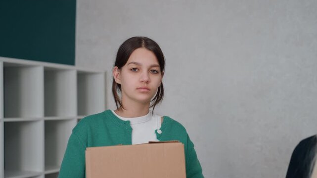 Young woman holding box in apartment, tense exchange with roommate, cardboard stacks and empty shelving, neutral interior, resigned expression, packing and relocation activity, candid indoor portrait,