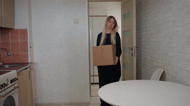 Woman unpacks box in compact kitchen movingin scene showing cardboard box placed on round table, blonde tenant opens tape, arranges kitchenware on countertop, sunlight through window, minimal