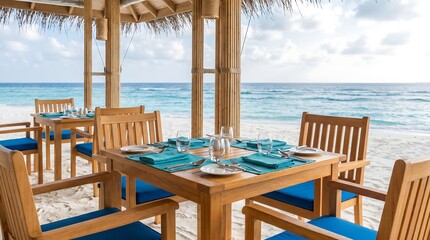 Fototapeta premium Empty wooden dining table with blue placemats and glasses in beachfront hut with thatched roof overlooking turquoise ocean and white sand beach in Maldives