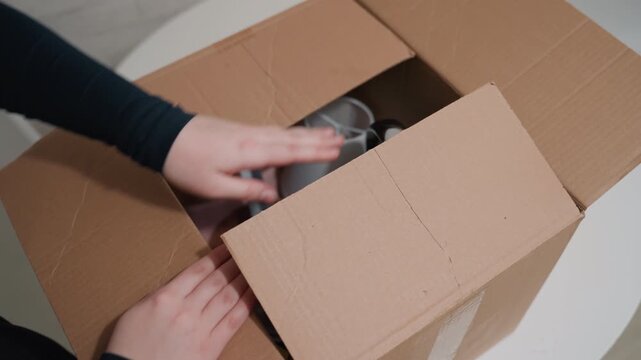 Packing kitchenware into cardboard box, hands placing kettle and mugs, closeup of cardboard flaps, careful arrangement, quiet moving day, small apartment table, new home preparation, fragile items