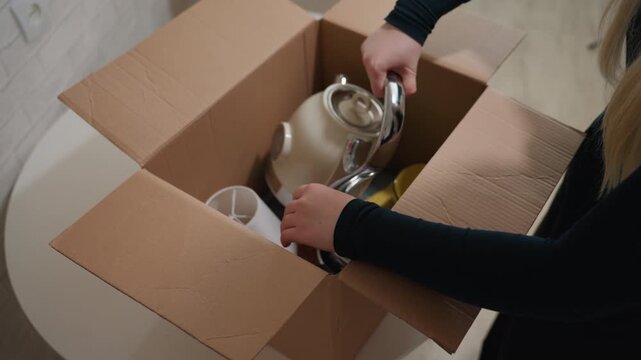 Woman packing teapot into cardboard box on round table, careful hands wrapping kettle and glass cup, soft natural light through window, minimalist apartment interior, moving day mood, organizing