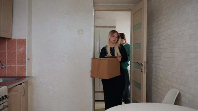 Friends unpack boxes in small kitchen, roommate lifts cardboard box into apartment, helper organizes utensils on round table, light conversation, warm cooperative mood, settling into new home