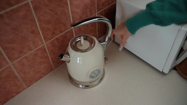 Kettle placed on base in kitchen, hand from green sweater sets kettle and presses switch, indicator light glows blue, tiled backsplash and microwave nearby, morning beverage preparation while settling