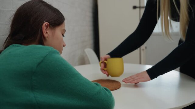 Girl at table receiving yellow mug, tender moving day scene in compact apartment caregiver hand places mug on coaster near greensweatered teenager, doorway and natural light visible, gentle comfort