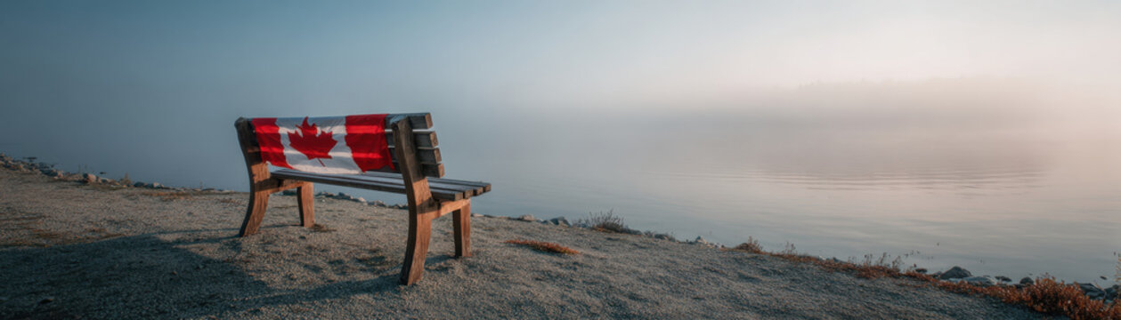 Wooden bench with red and white maple leaf flag overlooks calm lake on peaceful Canada day with soft morning light and clear sky