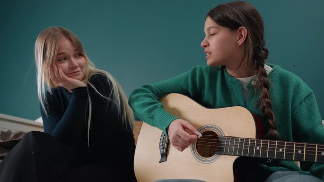 Two friends in new apartment playing acoustic guitar on floor, laughing and singing, braided hair, cozy knit sweater, teal wall background, casual unpacking with boxes, relaxed intimate jam session,