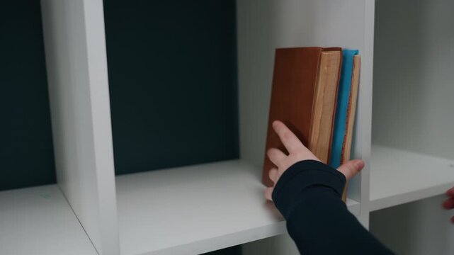 Hands arrange brown books then add terracotta plant on white cubby shelf with teal back close framing shows spines, stack placement and small succulent in clay pot, finishing touch of new apartment