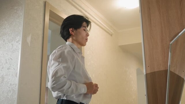 Young Asian businessman adjusting white shirt in dim hotel hallway, buttoning cuffs, wearing glasses with curly hair, tense but focused, preparing for job interview and later presentation, warm