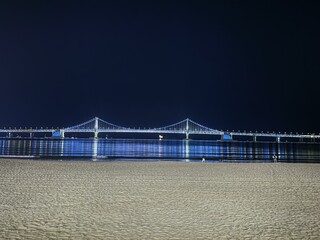 Fototapeta premium lluminated Suspension Bridge Glowing Blue at Night, Reflected on Calm Sea with Sandy Beach Foreground