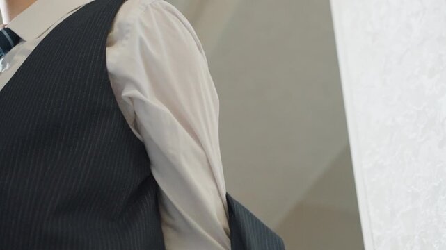 Man adjusting navy suit vest closeup, hands fastening buttons, straightening striped tie, tailored jacket over shoulder, soft indoor light, focused preparation for ceremony or meeting