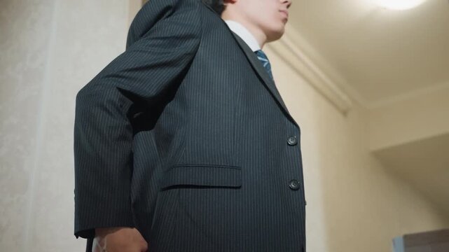 Young asian man putting on jacket in hallway foyer, stepping into light, straightening lapel and smoothing sleeve, confident posture preparing for interview or office meeting, cinematic lowangle