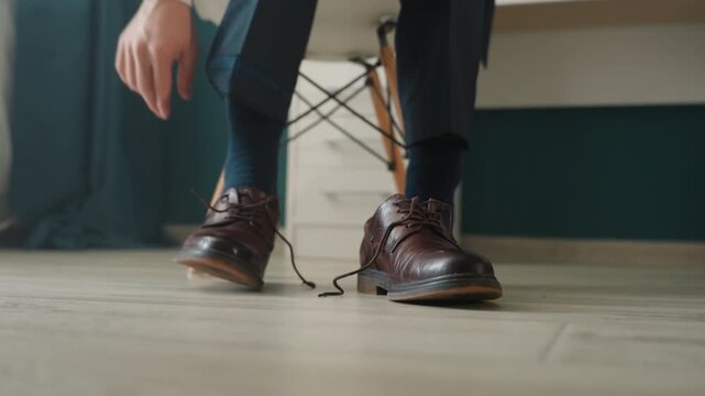 Polished brown leather shoes on floor man in suit sits on chair tying laces closeup hands show texture and shine cinematic minimalist mood meticulous preparation for interview or wedding