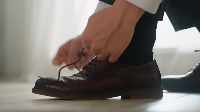 White man tying brown leather shoe, closeup hands smoothing sock and lacing brogue under soft sunrise light, suit trousers visible, careful executive preparing for wedding and meeting, composed mood,