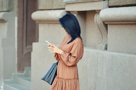 A woman in a flowing brown dress and a handbag stands against a stylish urban building. She is ordering a taxi online using the app in her smartphone
