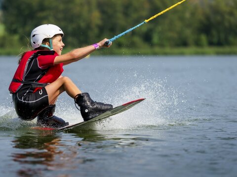 Boy or girl, sitting on wakeboard, cheerful, red life jacket, water skiing and summer holiday in wakepark, Str&aacute;ž pod Ralskem, Czech Republic