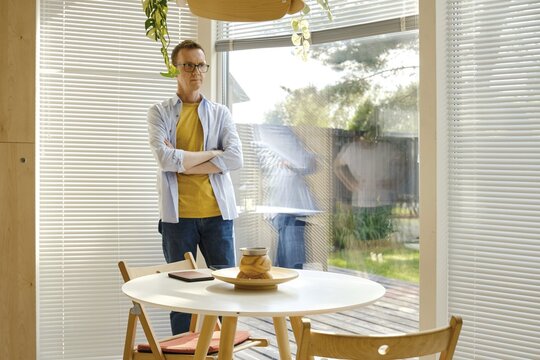A middle-aged man with glasses stands with crossed arms beside a round table in a well-lit, modern living area. The natural light illuminates the space, creating a calm atmosphere