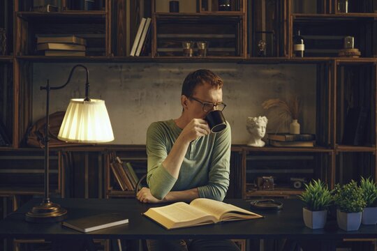 A middle-aged man sits at a desk in a cozy room, sipping a herbal tea from a mug. Soft light from a lamp illuminates the book he reads before going to bed