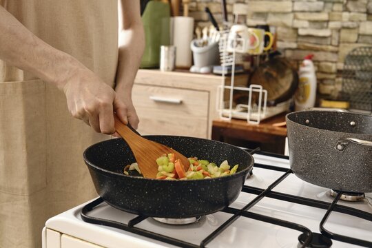 A person stirs a colorful mix of fresh vegetables in a frying pan on a stove in a cozy kitchen. Making vegetable base for ragout