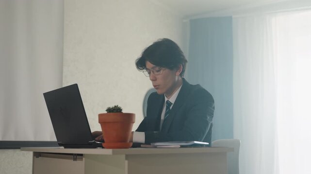 Businessman staring at laptop screen young professional in suit attending video call, potted plant foreground, ambient window light, thoughtful expression, taking notes, remote interview scenario,