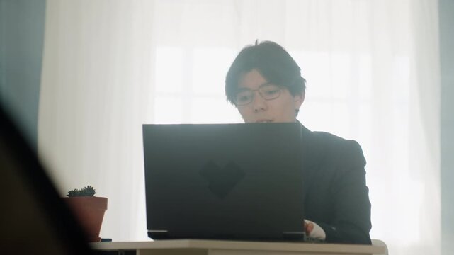 Asian man at laptop, soft backlight, quiet home office. Pensive remote developer reviewing code and documents, freelancer writing notes, consultant joining video call, glasses, small potted plant