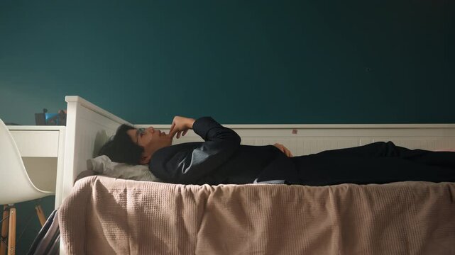 Young person on bed holding phone, intimate latenight conversation scene teal wall and soft lamp glow, side table and chair visible, rumpled blanket, profile turned upward, quiet apartment atmosphere,