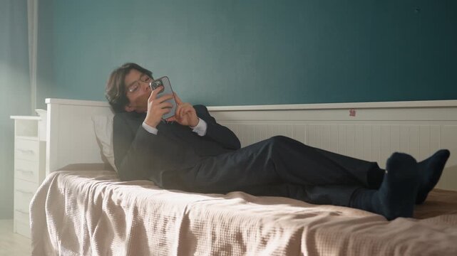 Asian professional lying on bed scrolling phone, wide frame shows suitclad full body, socks and linen, sunlight streaming from window, bedside drawer visible, relaxed posture and reflective gaze,