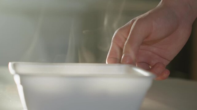 Hand testing hot steaming container on kitchen tabletop, closeup of fingers hovering over plastic tub, visible vapor rising, soft morning light, careful motion conveying warmth and safety