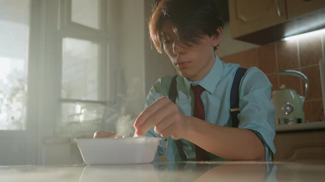 Young man eating breakfast in kitchen, soft golden sunlight through window, student in shirt and suspenders stirs steaming bowl of cereal with spoon, contemplative mood, warm vintage tones and cozy