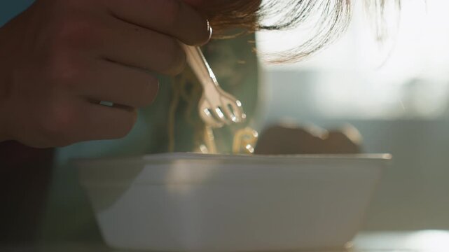 Closeup hands eating noodles in sunlight, backlit foam container on table, plastic fork twirling steaming strands, quiet morning routine, intimate comfort and warm tones capturing candid home moment.
