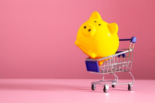 Yellow piggy bank in shopping cart on pink background. Save money for future concept.