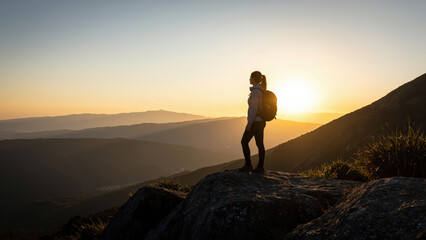 Obraz premium Summit Serenity: A lone hiker, silhouetted against a breathtaking sunset, stands on a rocky peak, gazing out at the vast, layered mountain landscape, embodying solitude and the spirit of adventure.