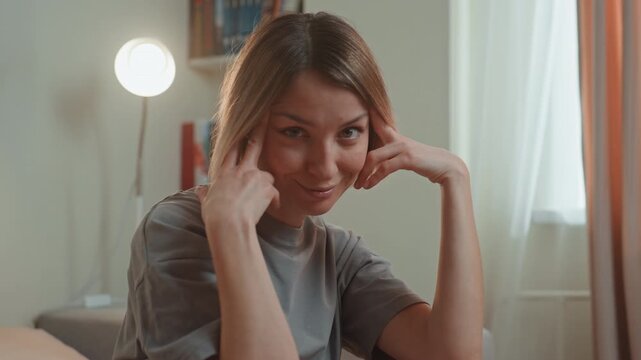 Young woman at home listening to music, smiling and miming headphones, tapping temples in softly lit living room with lamp and bookshelf, casual gray shirt, playful lover vibe, intimate cozy