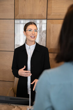 Adult Caucasian female receptionist assisting to guest at hotel reception, explaining check-in procedure, registration details at front desk. Hospitality, professional service, travel industry concept