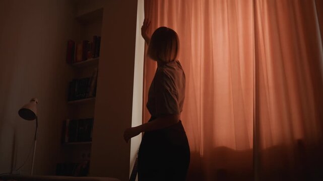 Woman opening coral curtains at dawn. Silhouette sequence inside quiet bedroom short blonde woman stands in shadow, draws thick drapery to reveal soft morning rays, stretches arms upward, peers