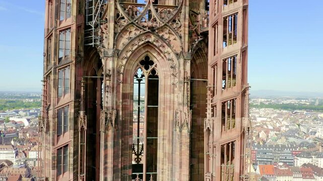 Strasbourg, France. The soaring Gothic spire of Notre-Dame Cathedral, captured in a continuous aerial ascent revealing intricate pink sandstone tracery and the cityscape below under a clear blue sky.