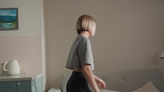 Woman playfully fluffing pillow beside gray couch and teal cabinet, bright neutral palette, kettle and lamp on side table, energetic gesture and casual style suggesting music mood and lighthearted