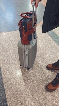 Individual with brown shoes holds suitcase with a bag on top, standing on polished floor in a busy airport terminal, showcasing travel essentials and personal belongings